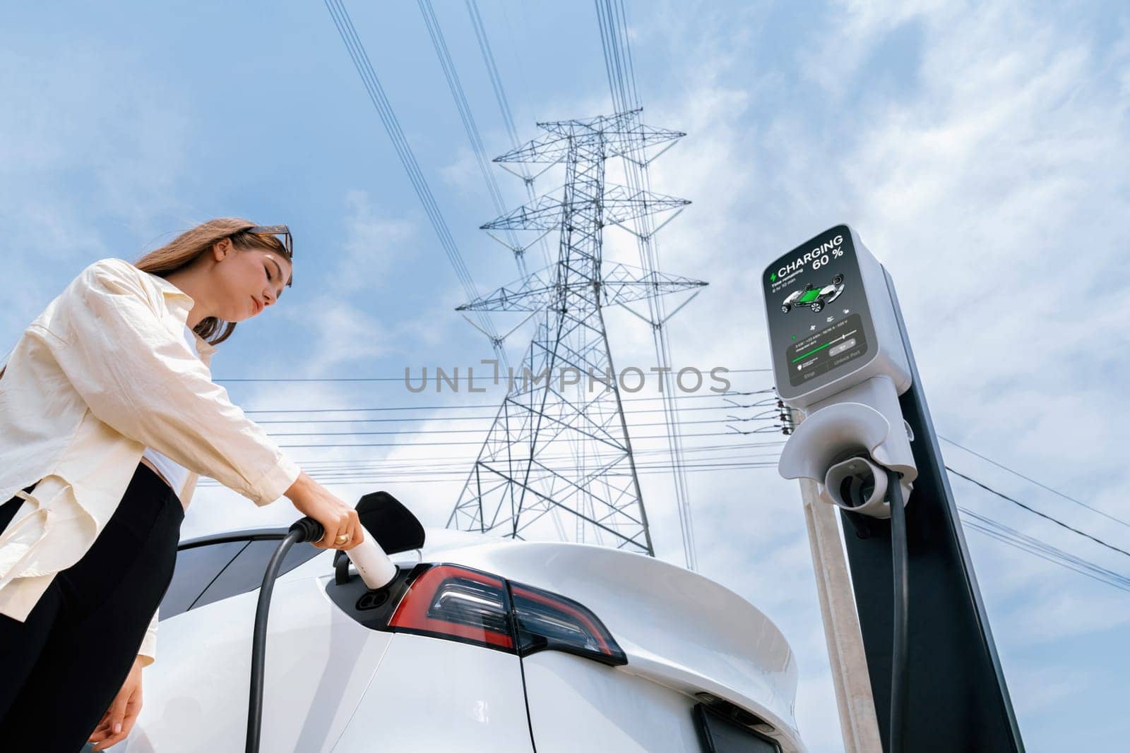 Woman recharge EV electric car battery at charging station connected to electrical power grid tower on sky background as electrical industry for eco friendly vehicle utilization. Expedient