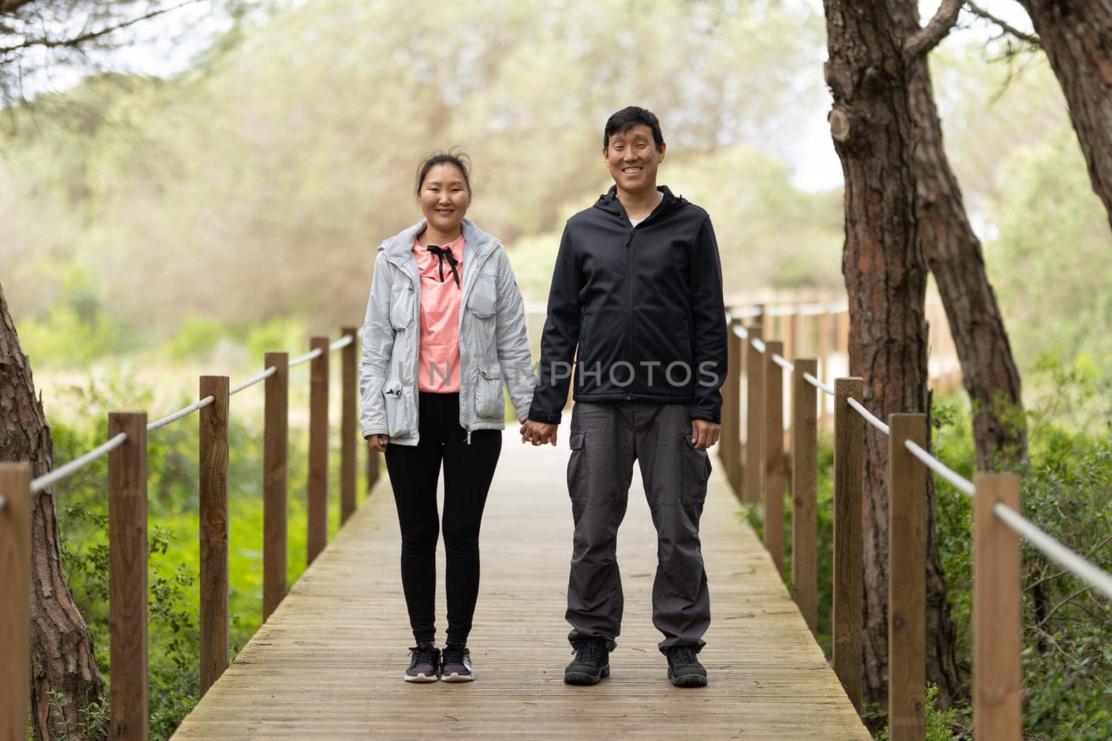 A man and woman are standing on a wooden bridge, holding hands. Scene is warm and affectionate, as the couple shares a moment together