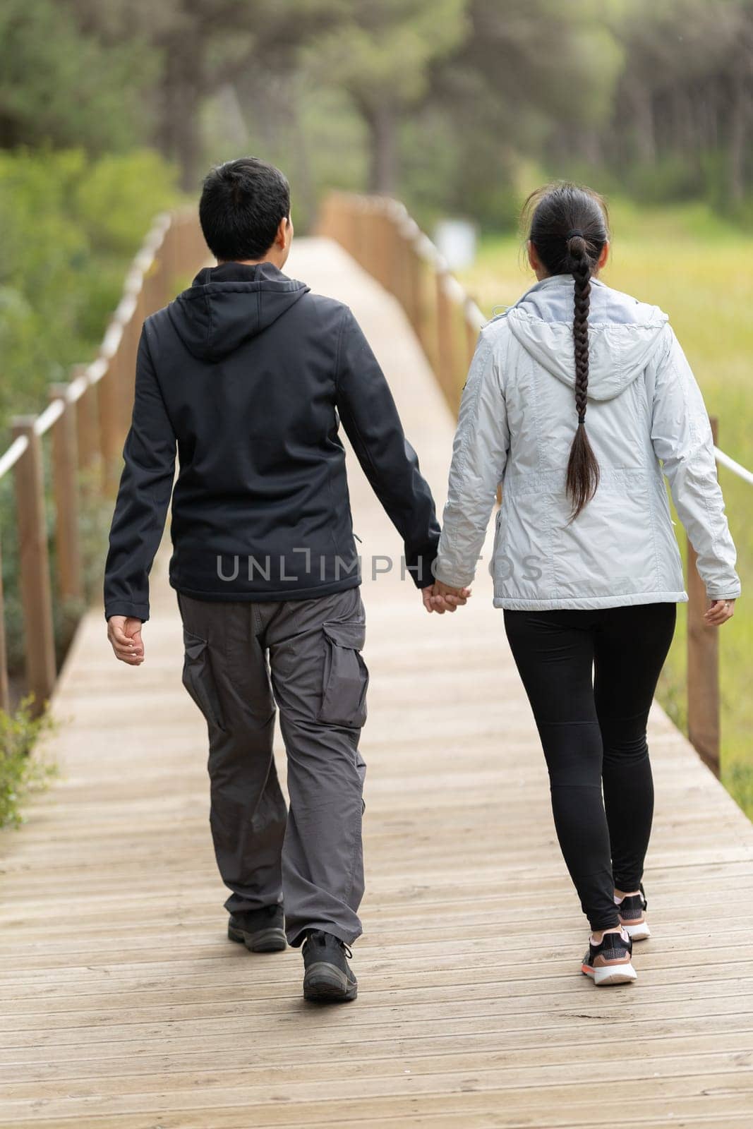 A man and woman are walking on a wooden bridge. The man is wearing a black jacket and the woman is wearing a gray jacket. They are holding hands and walking together