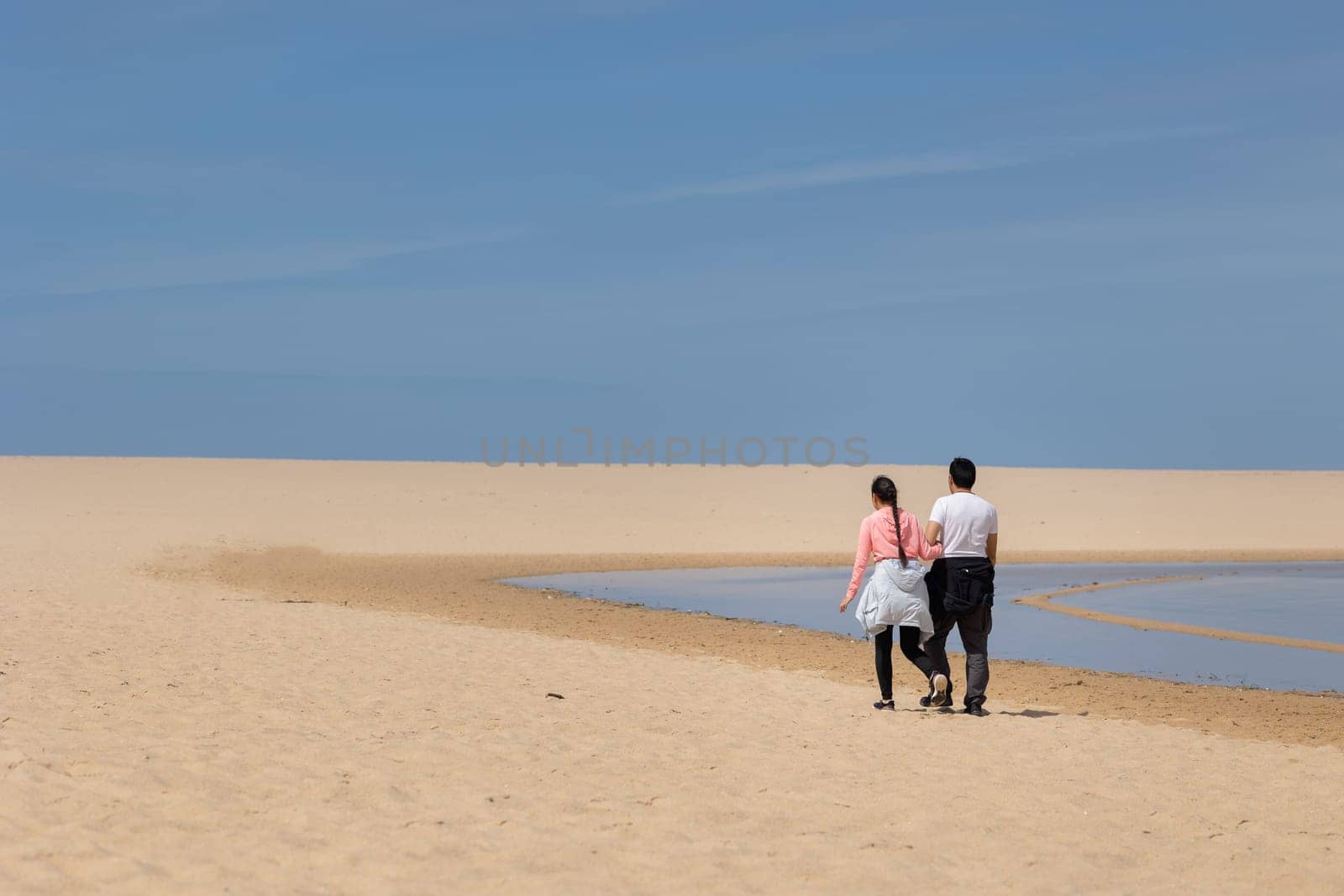 A couple walking on a beach with a clear blue sky above them. The beach is empty and the couple is the only ones there
