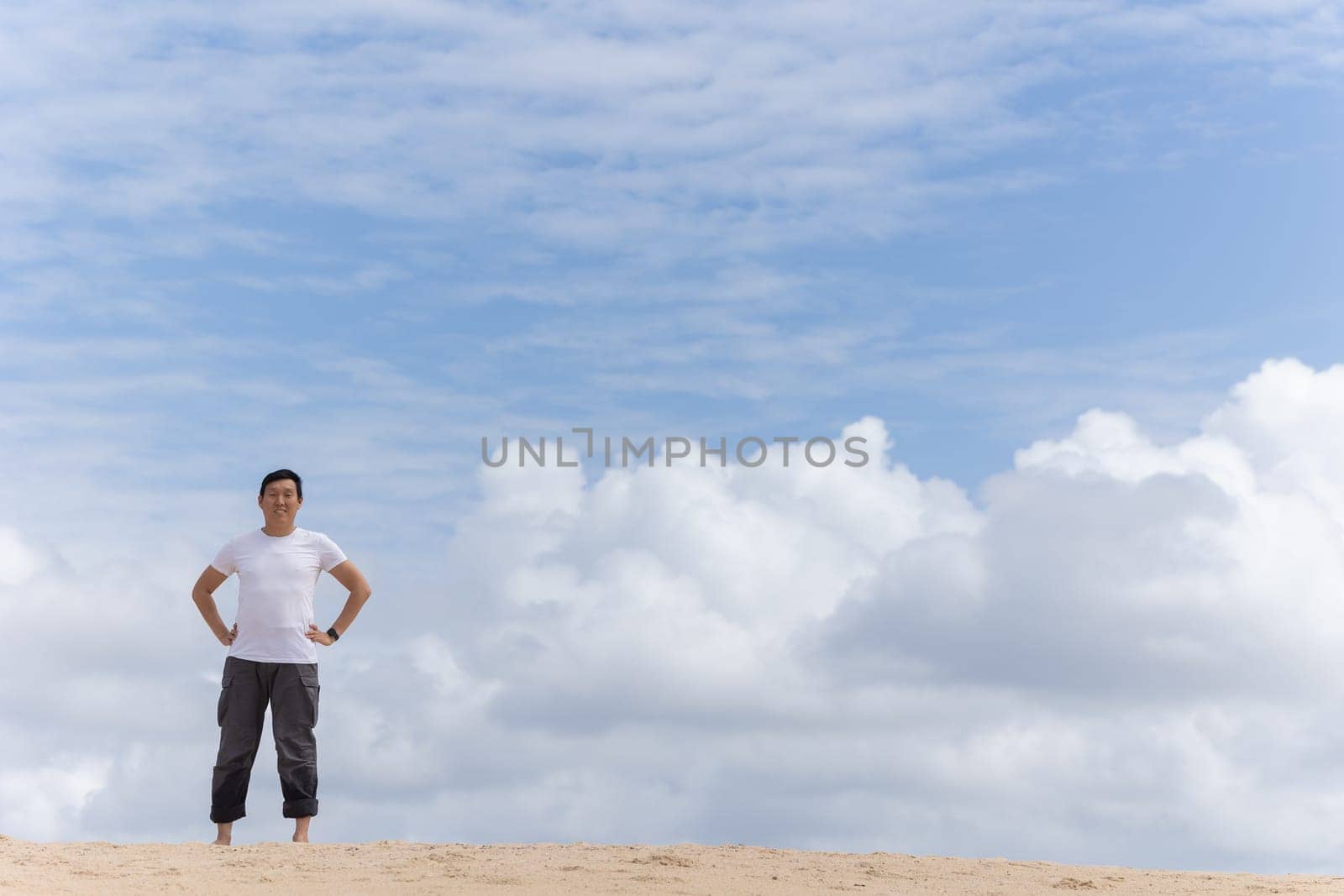 A woman stands on a beach with a cloudy sky in the background. She is wearing a white shirt and grey pants