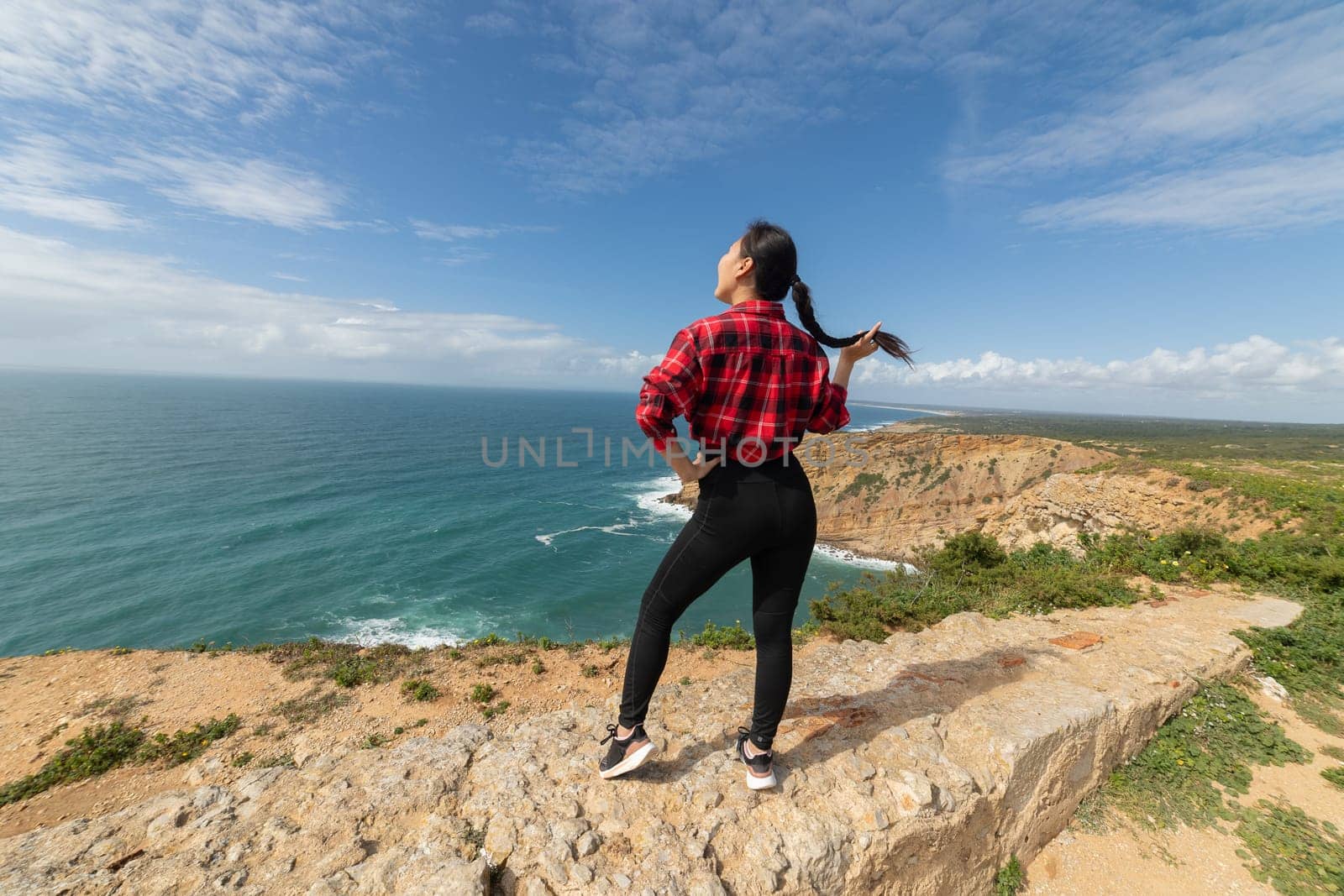 A woman stands on a rocky cliff overlooking the ocean. She is wearing a red and black plaid shirt and black pants. The sky is clear and blue, and the ocean is calm. The woman is enjoying the view