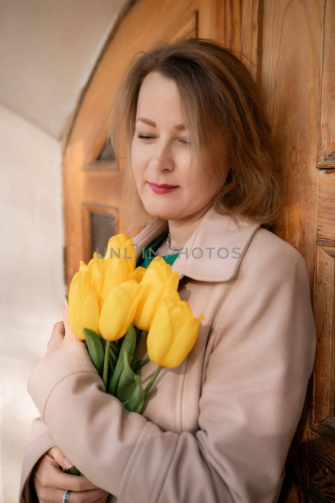 A woman is holding a bouquet of yellow tulips. She is wearing a tan coat and a green shirt