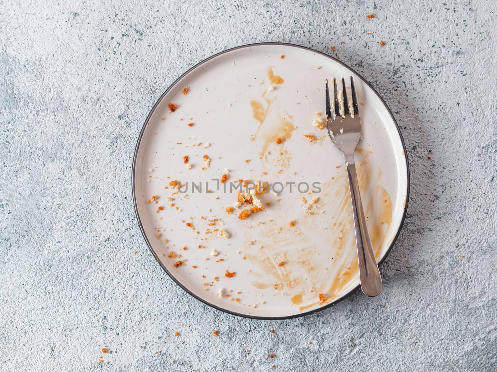Empty dirty dish after cheesecake with caramel sauce and dessert fork. White rustic trendy modern fashionable dirty plate on gray cement background, top view or flat lay. Copy space for text