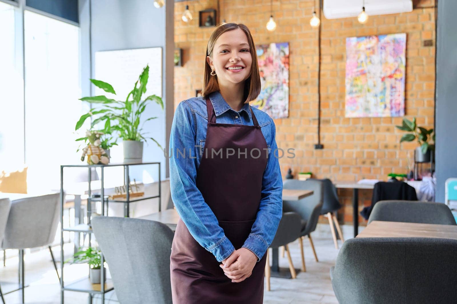 Young woman worker, owner in apron looking at camera in restaurant, coffee shop by VH-studio