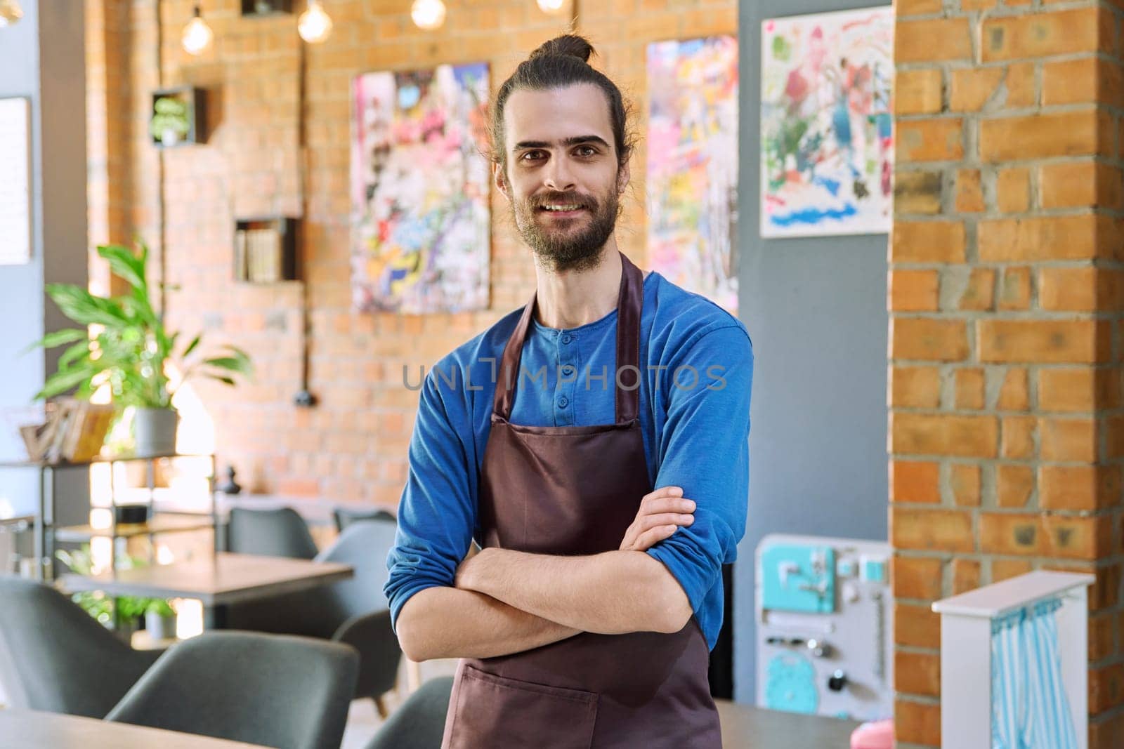 Confident successful young man service worker owner in apron with crossed arms looking at camera in restaurant cafeteria coffee pastry shop interior. Small business staff occupation entrepreneur work
