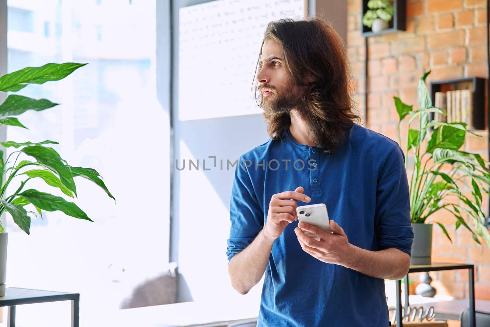 Young 30s stylish handsome serious bearded man with long hair using smartphone. Mobile technologies network Internet applications apps for leisure work business communication chatting education