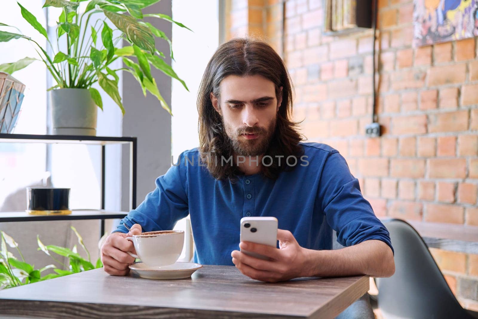 Young 30s stylish handsome bearded with long hair man using smartphone, drinking cup of coffee, sitting in cafe. Mobile technologies Internet apps applications for leisure work business communication