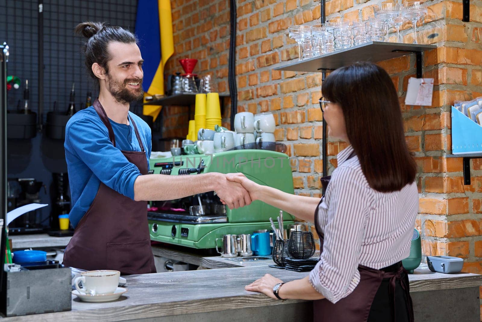 Young man in apron shaking hands with woman owner of coffee shop cafeteria restaurant, colleagues partners working together. Cooperation, small business, partnership, teamwork team, staff, success