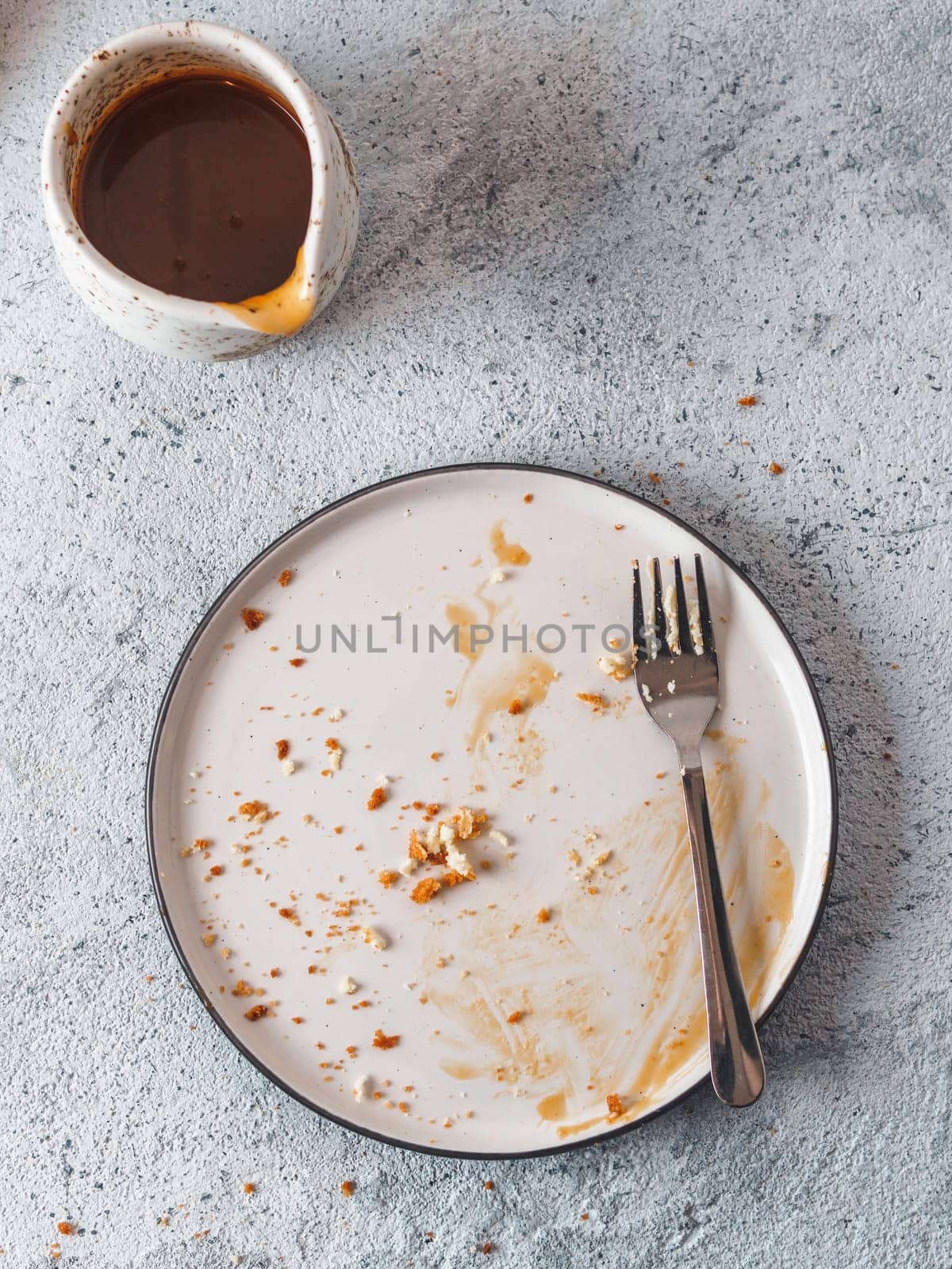 Empty dirty dish after cheesecake with dessert fork. Caramel sauce in sauceboat. White rustic trendy modern fashionable dirty plate on gray cement background, top view or flat lay. Copy space for text