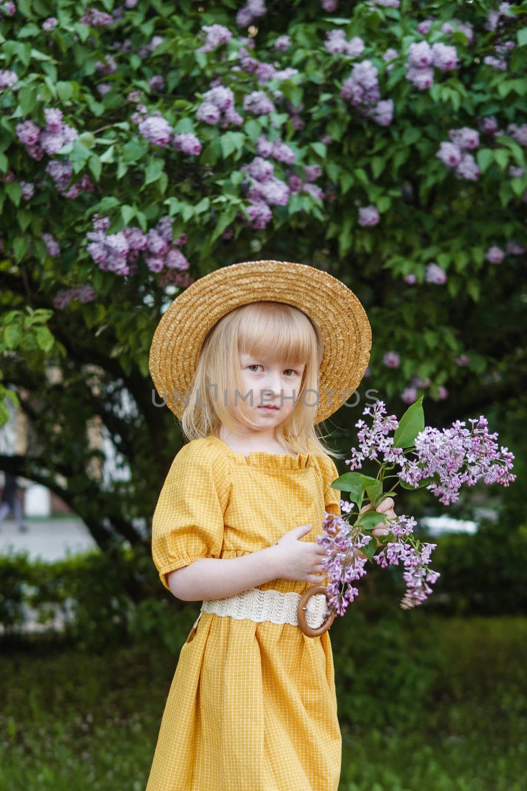 A little girl in a yellow dress and straw hat wearing a bouquet of lilacs. A walk in a spring park, blossoming lilacs