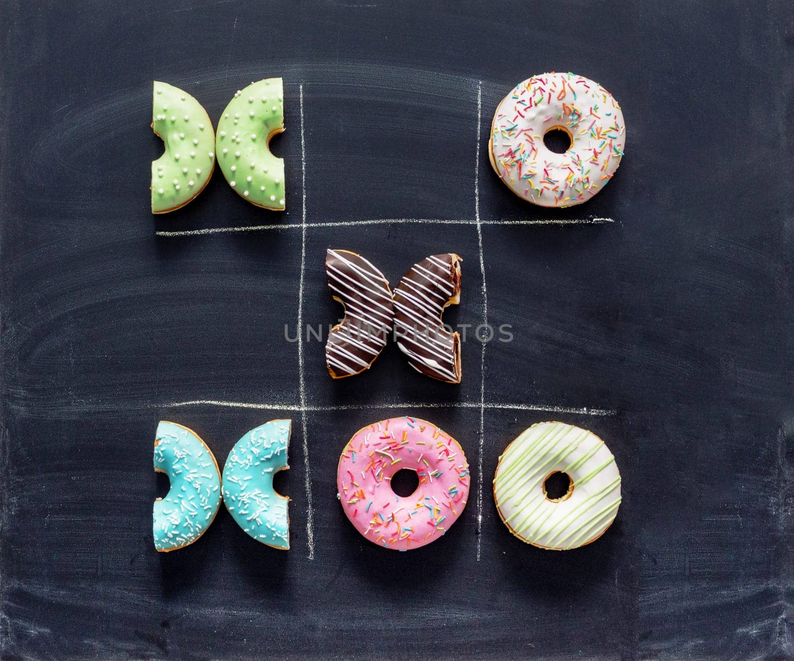 Tic tac toe made of donuts. Game noughts and crosses with donuts on chalkboard. Unhealthy diet, diabetes, sugar concept. Top view or flat lay.