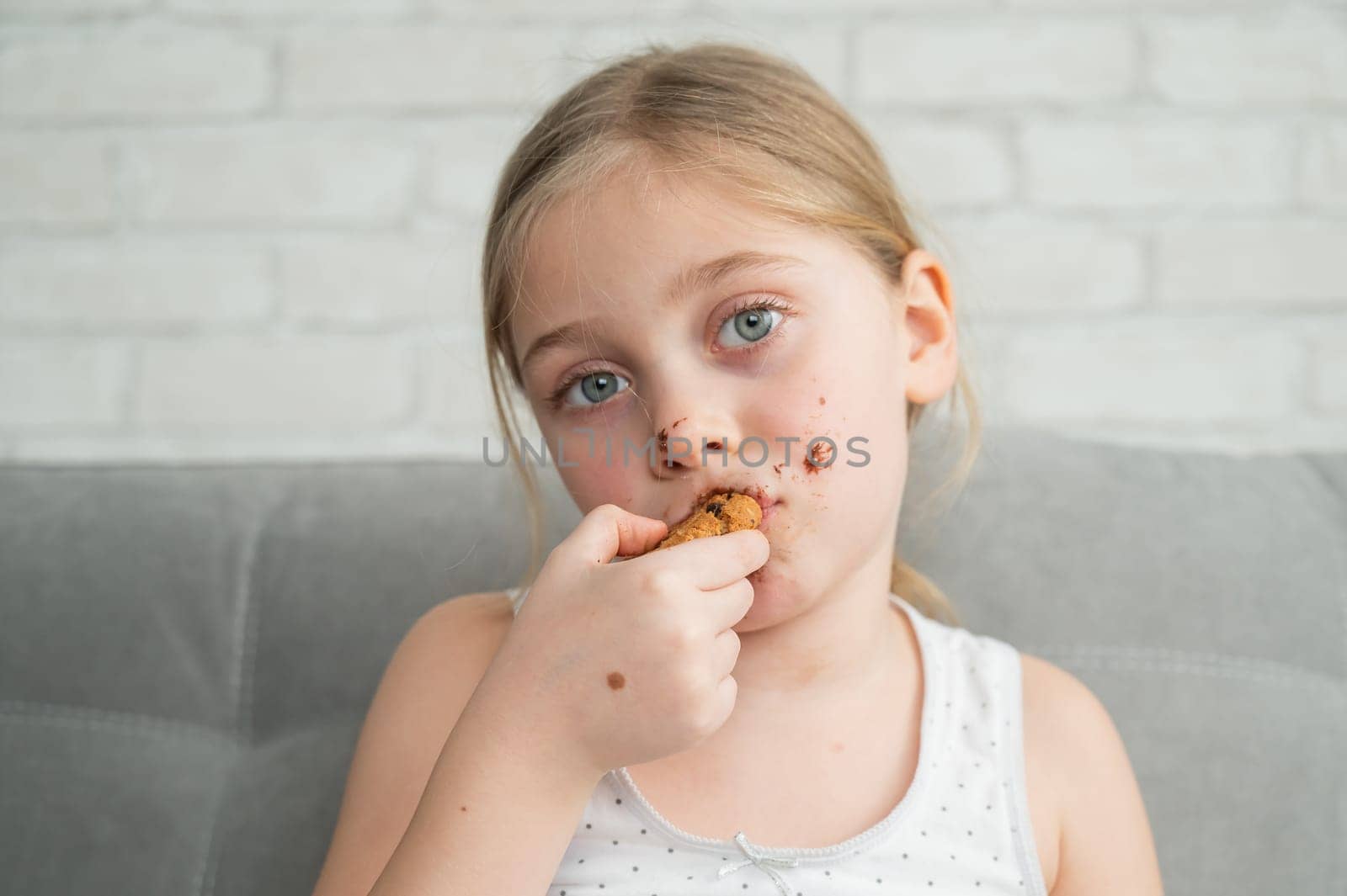 A cute little girl covered in chocolate eats cookies while sitting on the sofa