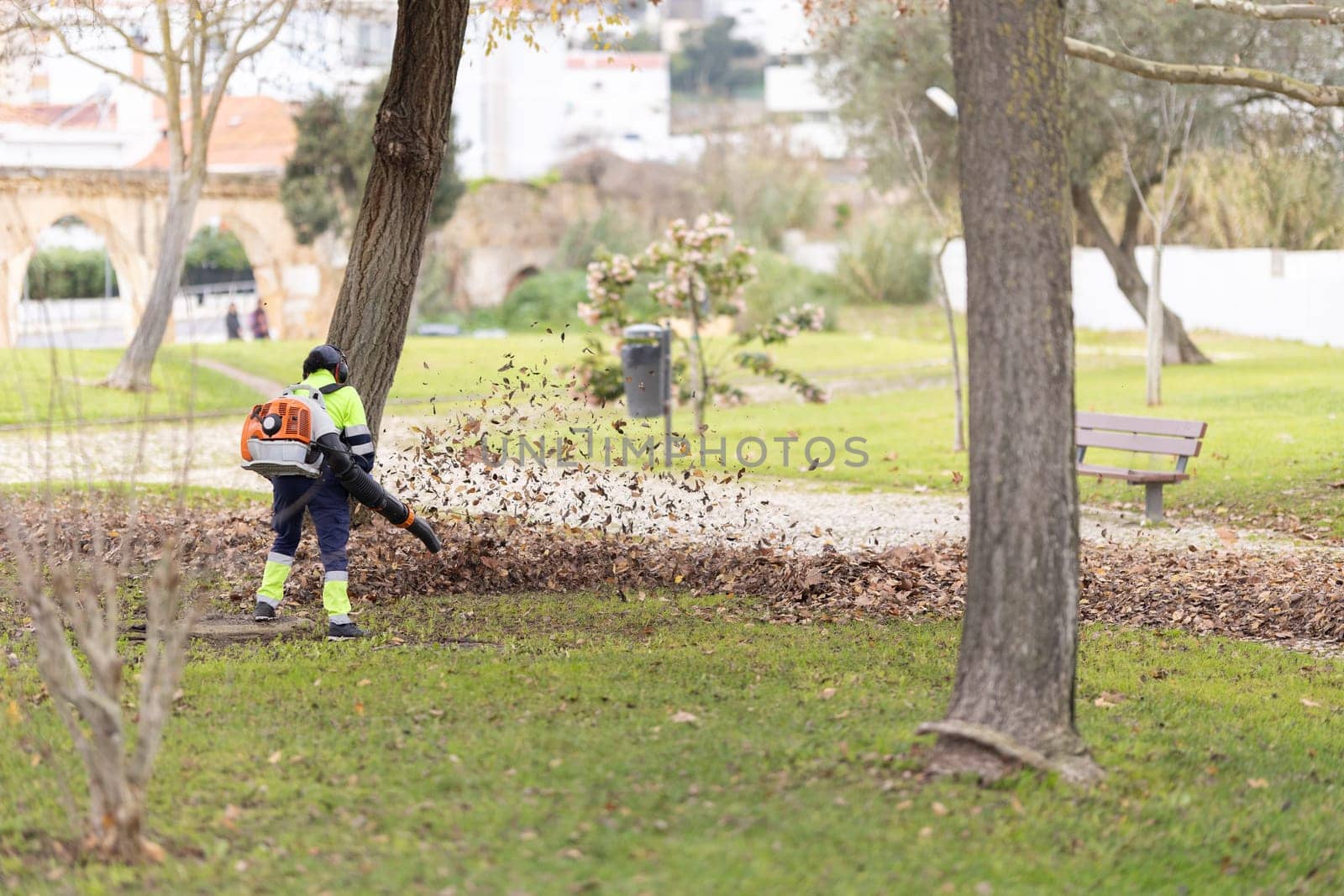 A wirker is blowing leaves with a leaf blower in a park. The leaves are scattered all over the ground, and the man is working hard to clear them. The scene is peaceful and serene