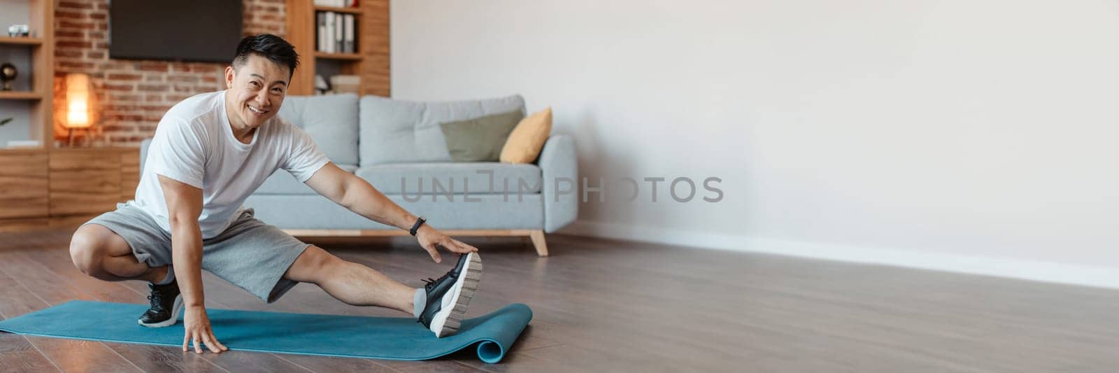 Happy asian mature man training at home, stretching legs in living room interior, panorama with free space. Positive middle aged guy exercising at home, enjoying healthy lifestyle