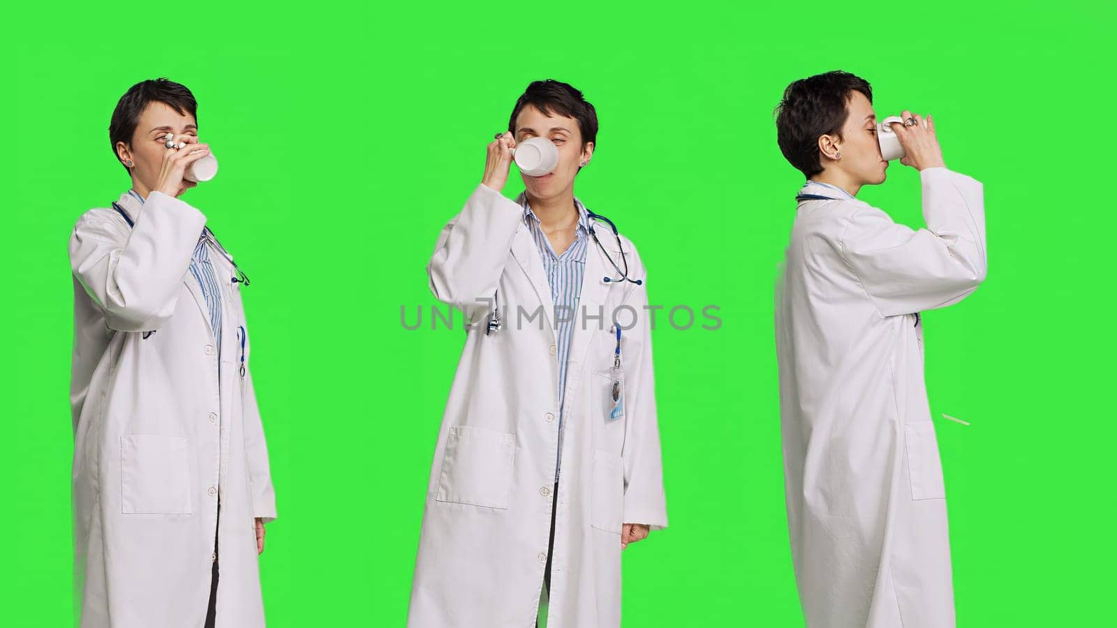 Woman physician drinking a cup of coffee against greenscreen backdrop, taking a break from medical work. Specialist serving caffeine refreshment in studio, wearing a white coat. Camera B.