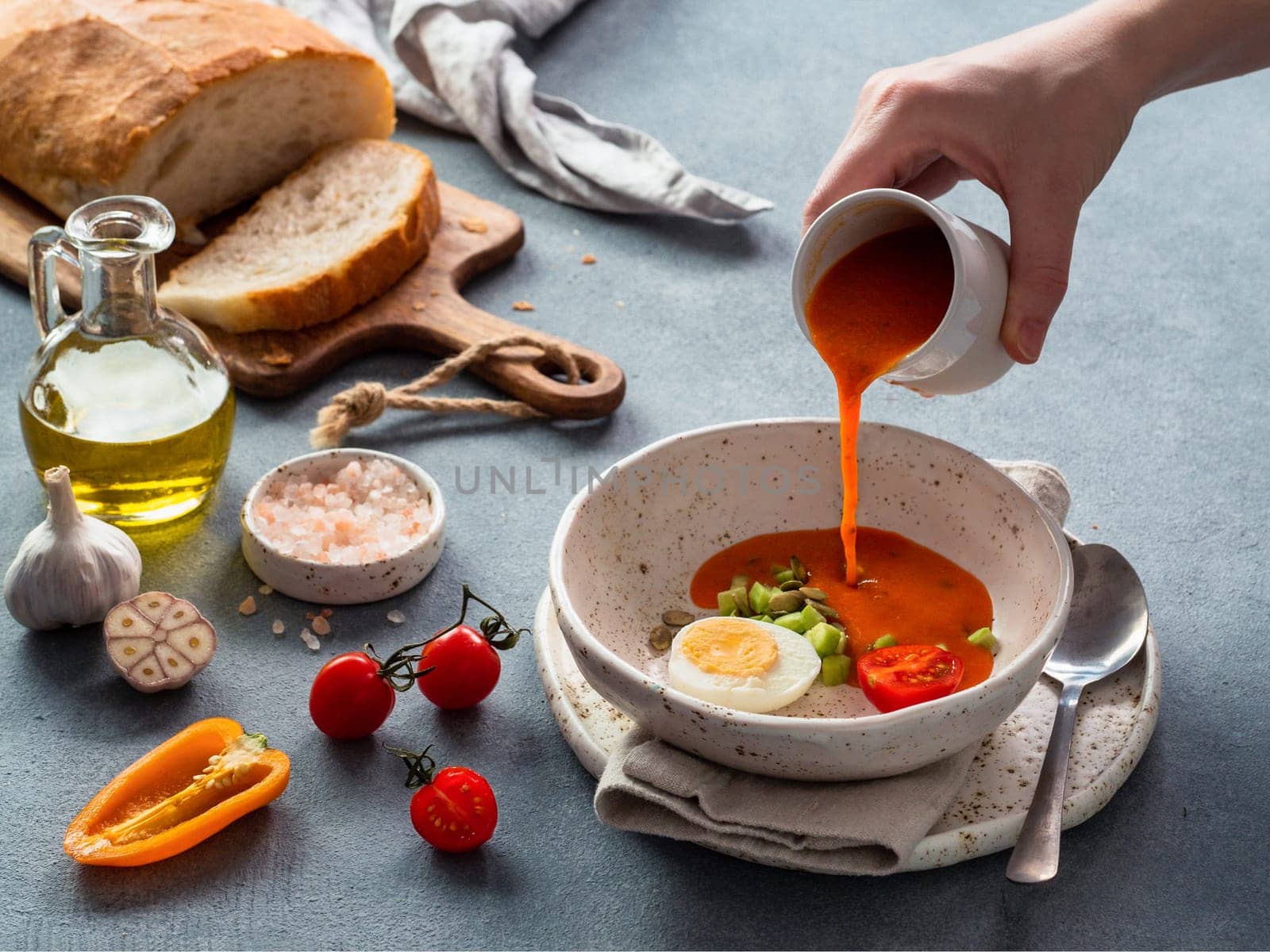 Gaspacho soup and ingredients. Woman's hand pouring of traditional spanish cold soup puree gazpacho in bowl on gray background.
