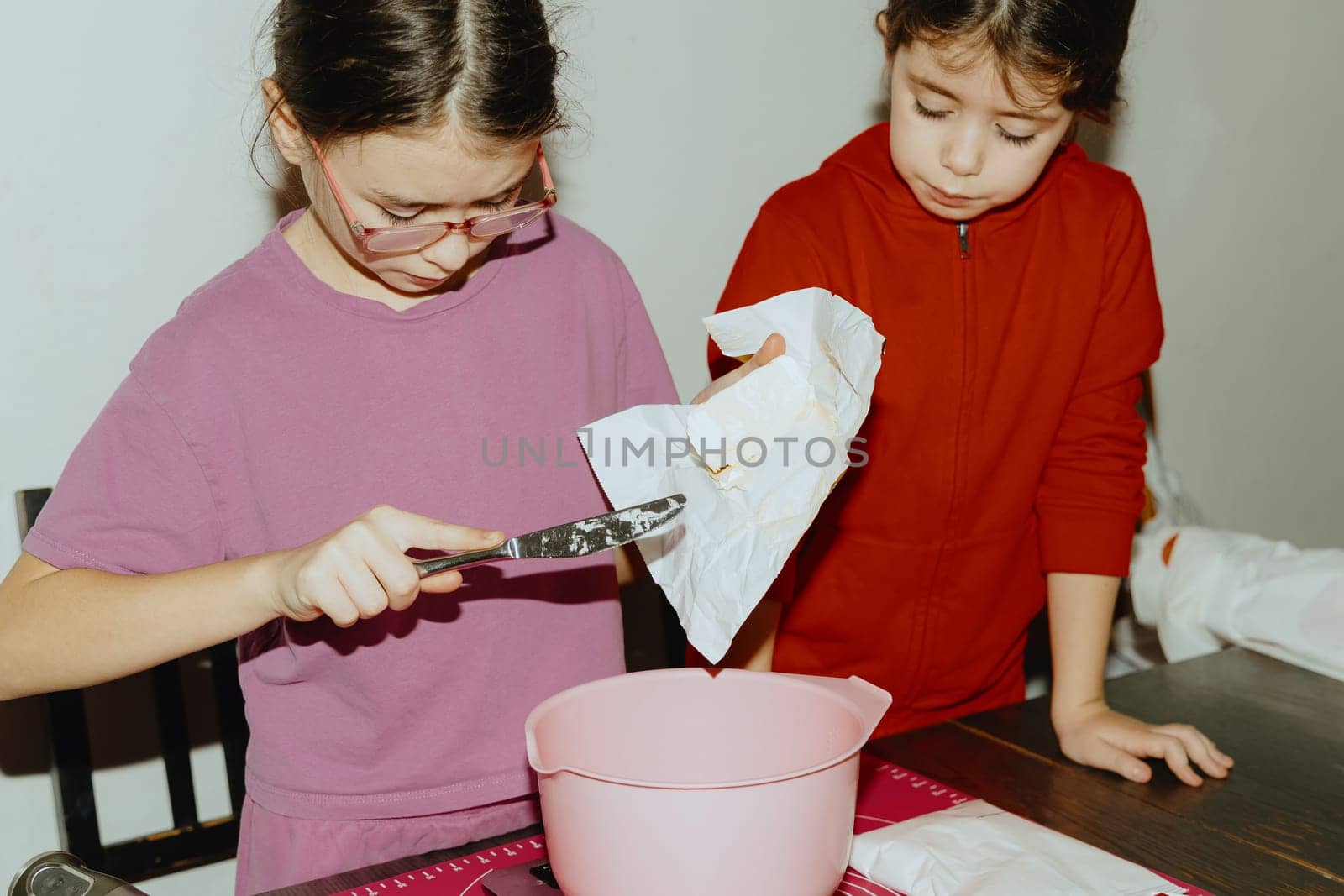 Two beautiful Caucasian brunette girls are standing at a table with food and one is cutting a piece of butter into a bowl for baking cookies, standing at the table in the kitchen during the day, close-up side view. Step-by-step instructions. Step 2.