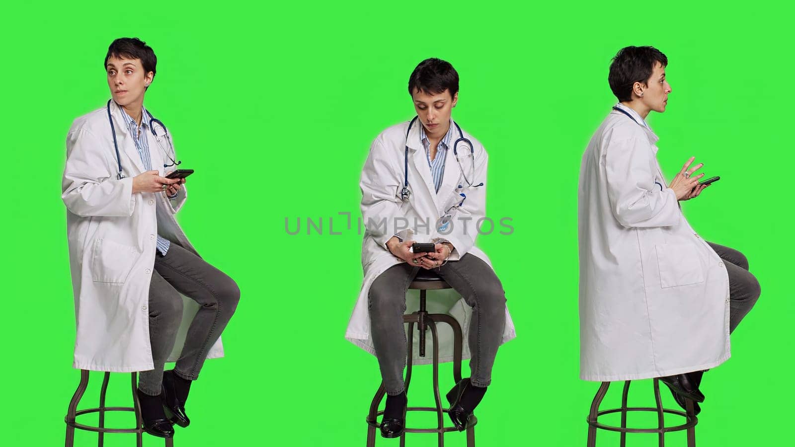 Woman specialist using smartphone social media apps in studio, sitting on a chair against greenscreen backdrop. Physician waiting for someone and browses online webpages, texting. Camera A.