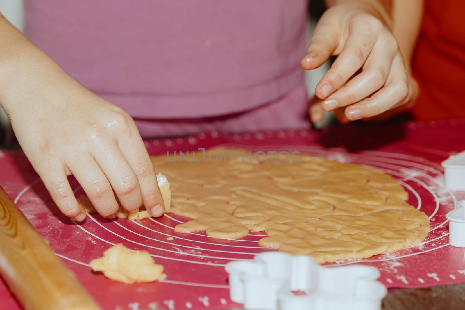 One unrecognizable Caucasian girl stands at the table and removes excess cut yellow shortbread dough lying on a pink silicone mat, close-up side view. Step by step instructions. Step 15