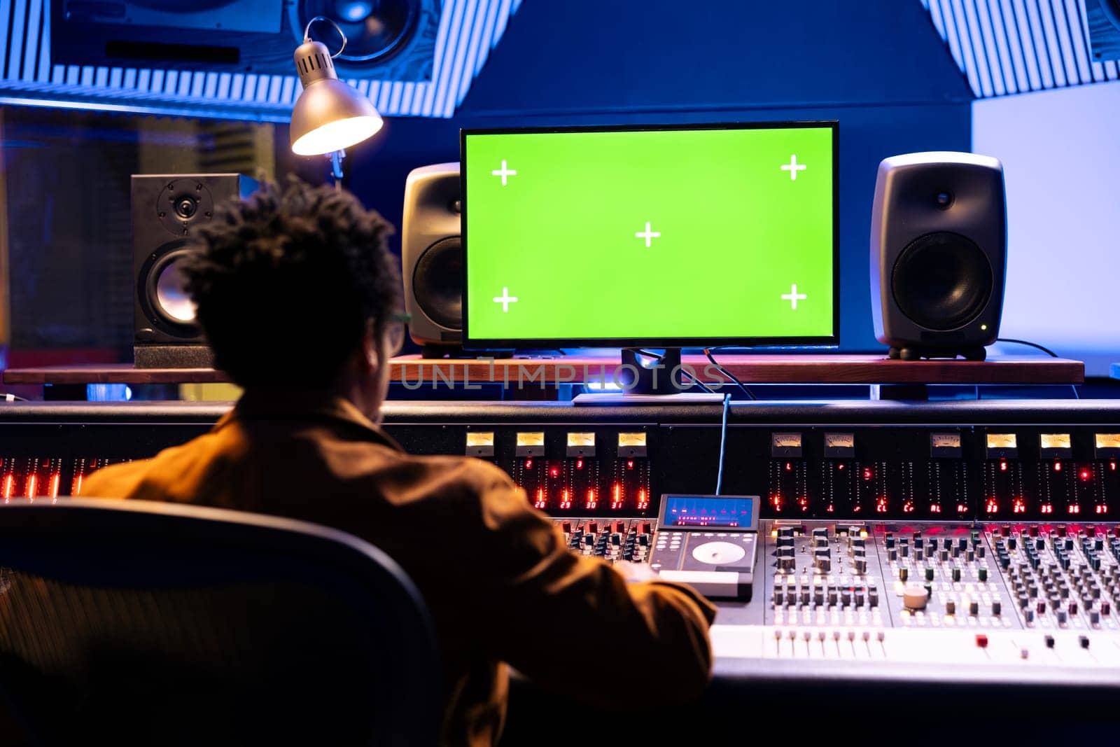 African american sound engineer works with greenscreen on computer in professional recording studio, mixing and mastering sounds on panel board. Young music producer pushing knobs and faders.