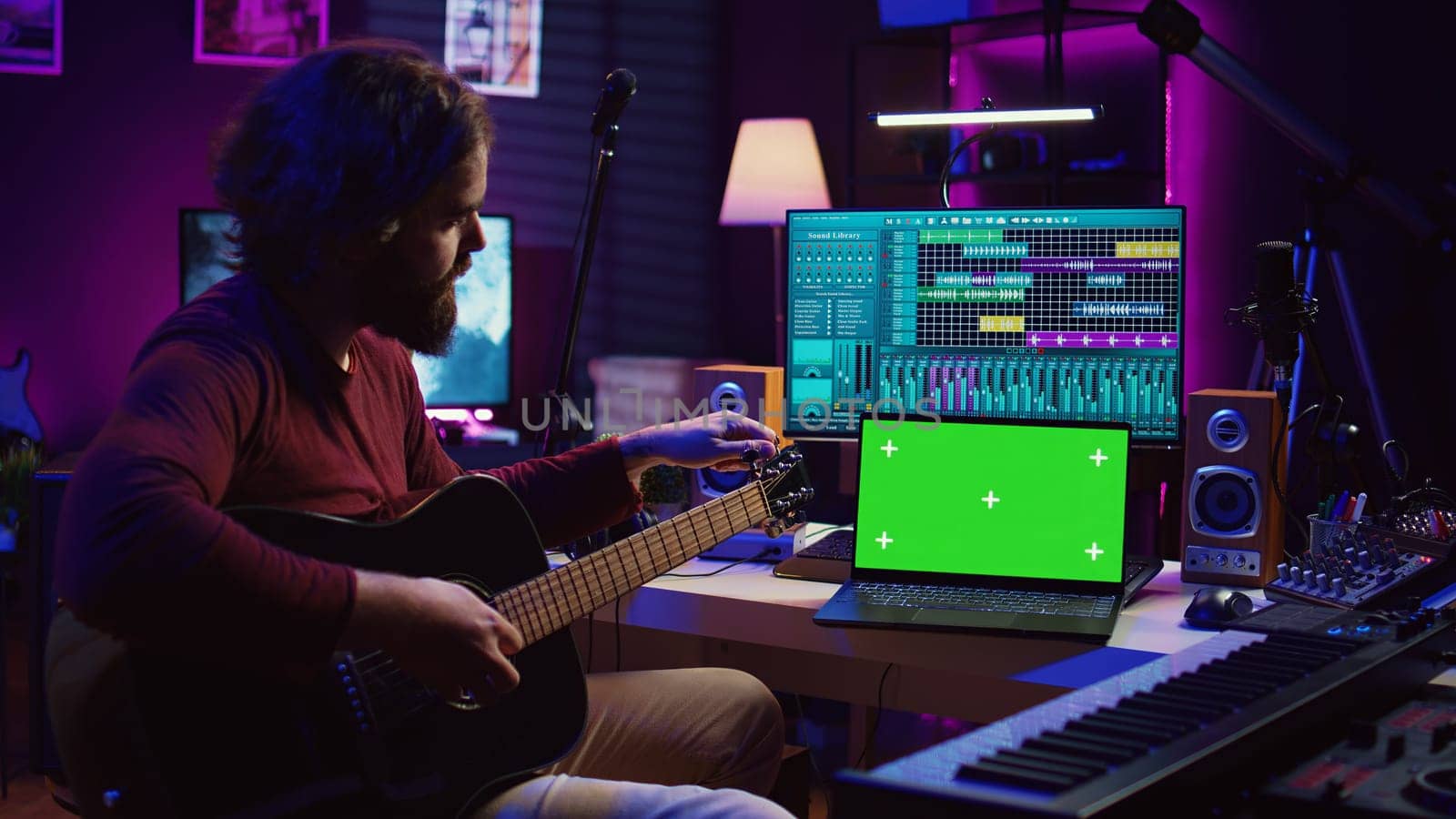 Musician learning to play new acoustic instrument in his home studio, practicing online guitar lessons on isolated greenscreen display. Watching tutorial on laptop to develop his skills. Camera B.