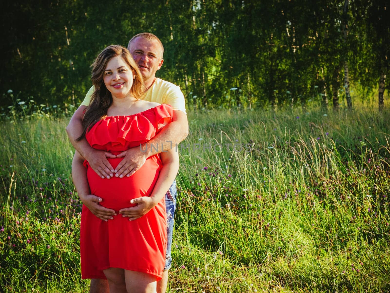Smiling beautiful pregnant woman in red dress and her husband with hands on belly outdoors. Man embraces from behind belly pregnant wife and smiling