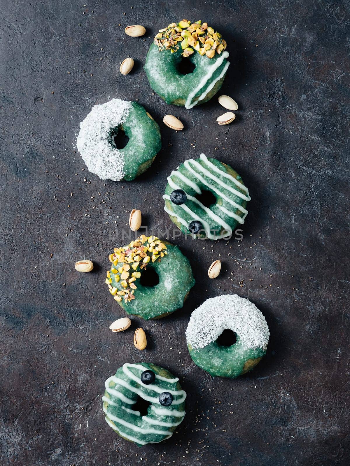 Vegan doughnuts with chia seeds topped with healthy spirulina glaze with pistachio, desiccated coconut and blueberry. Blue green spirulina donuts, on black background, top view or flat lay. Vertical