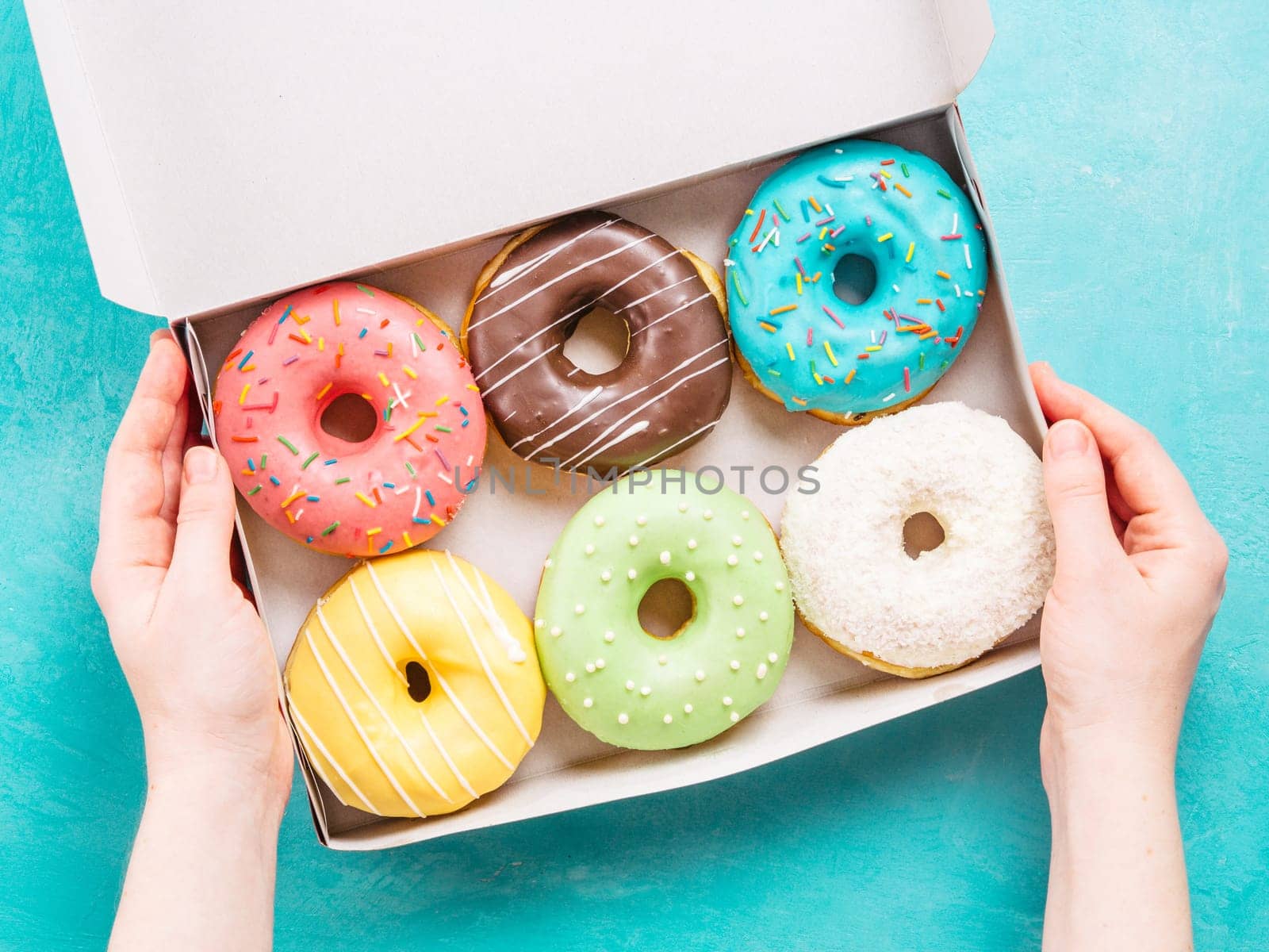 Top view of female hands holding box with colorful donuts on blue concrete background