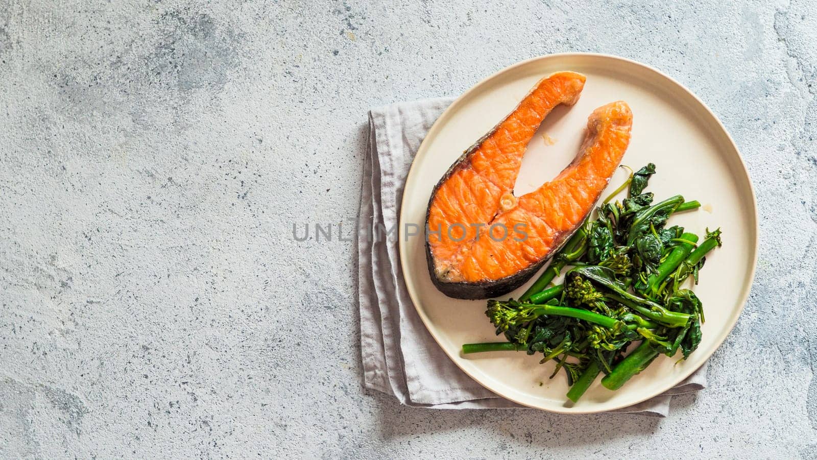 Ready-to-eat grilled salmon steak and greens - baby broccoli or broccolini and spinach on rustic craft plate over gray background. Keto diet dish. Top view or flat lay.Copy space left for text. Banner