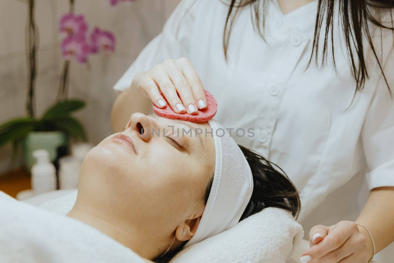 A girl cosmetologist cleanses the patient s face with a scrubbing sponge. by Nataliya
