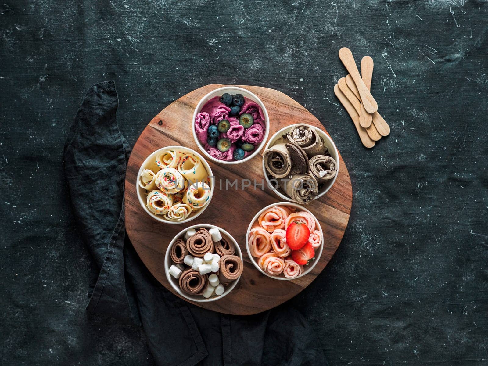 Rolled ice cream in cone cups on rustic round wooden tray on dark background. Different iced rolls top view or flat lay. Thai style rolled ice cream