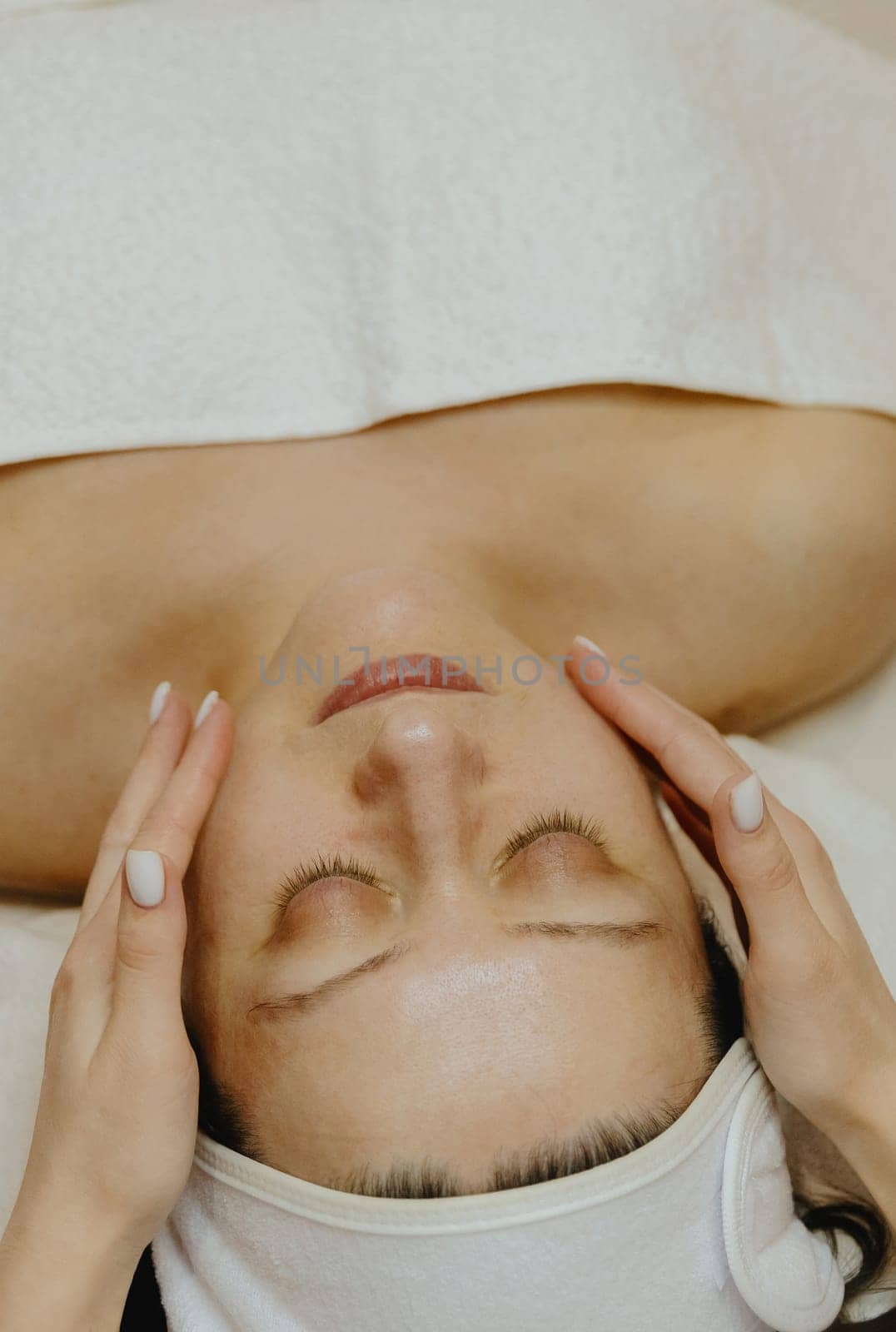 A young unrecognizable girl cosmetologist gently applies moisturizing cream with her fingers in the cheek area with stroking movements on the face of an adult client, who lies with her eyes closed on a massage table in a beauty salon, close-up side view.