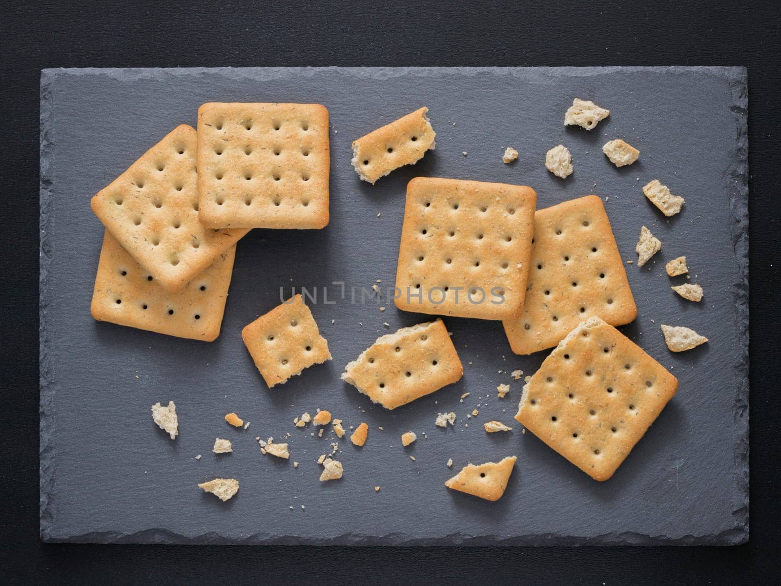 Square crackers with pieces and crumbs on slate gray background. Dry salt cracker cookies with fiber and dry spices. Top view or flat lay.