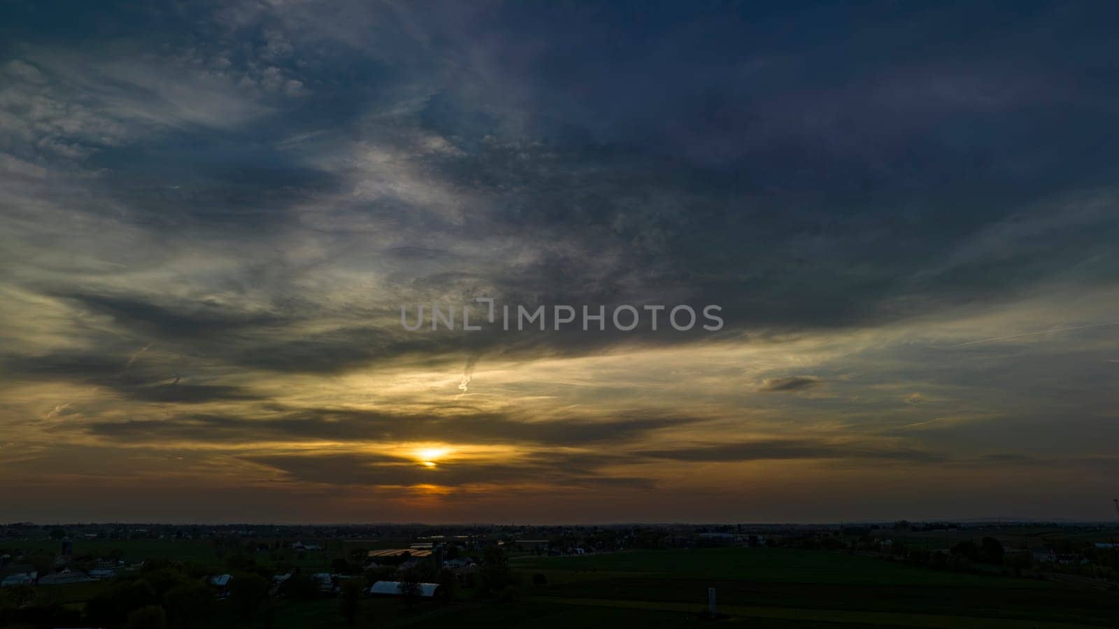 Dramatic Sunset Casting A Warm Glow Over A Rural Landscape With Silhouetted Trees And Buildings Under A Cloud-Streaked Sky.