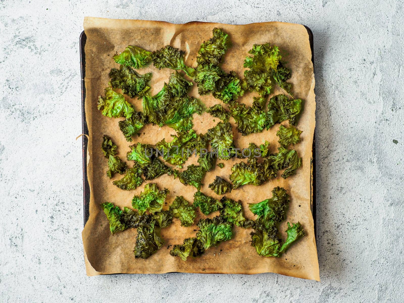 Green Kale Chips with salt on oven-tray. Homemade healthy snack for low carb, keto, low calorie diet. Gray cement background. Ready-to-eat kale chips, top view or flat lay.