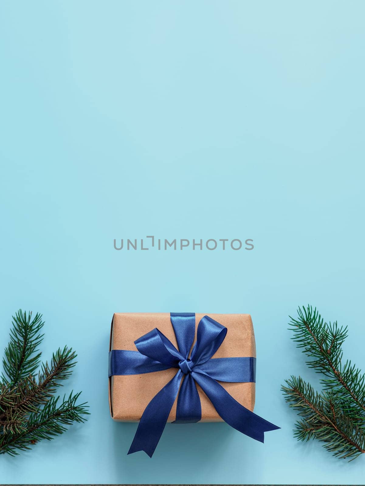 Gift box in craft wrapping paper and blue satin ribbon on turquoise blue background, copy space top. Beautiful Christmas or New Year present and fir branches, flat lay or top view. Vertical