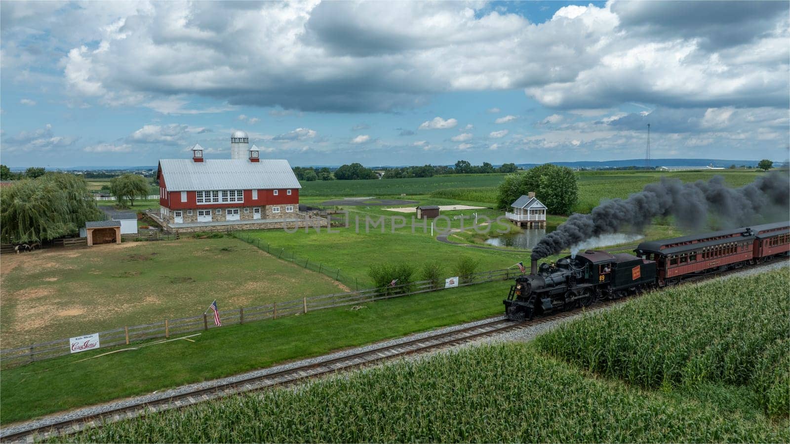 Ronks, Pennsylvania, August 15, 2023 - A vintage locomotive steams by a picturesque farm with its red barn and pond, adding a touch of historical charm to the rural landscape.