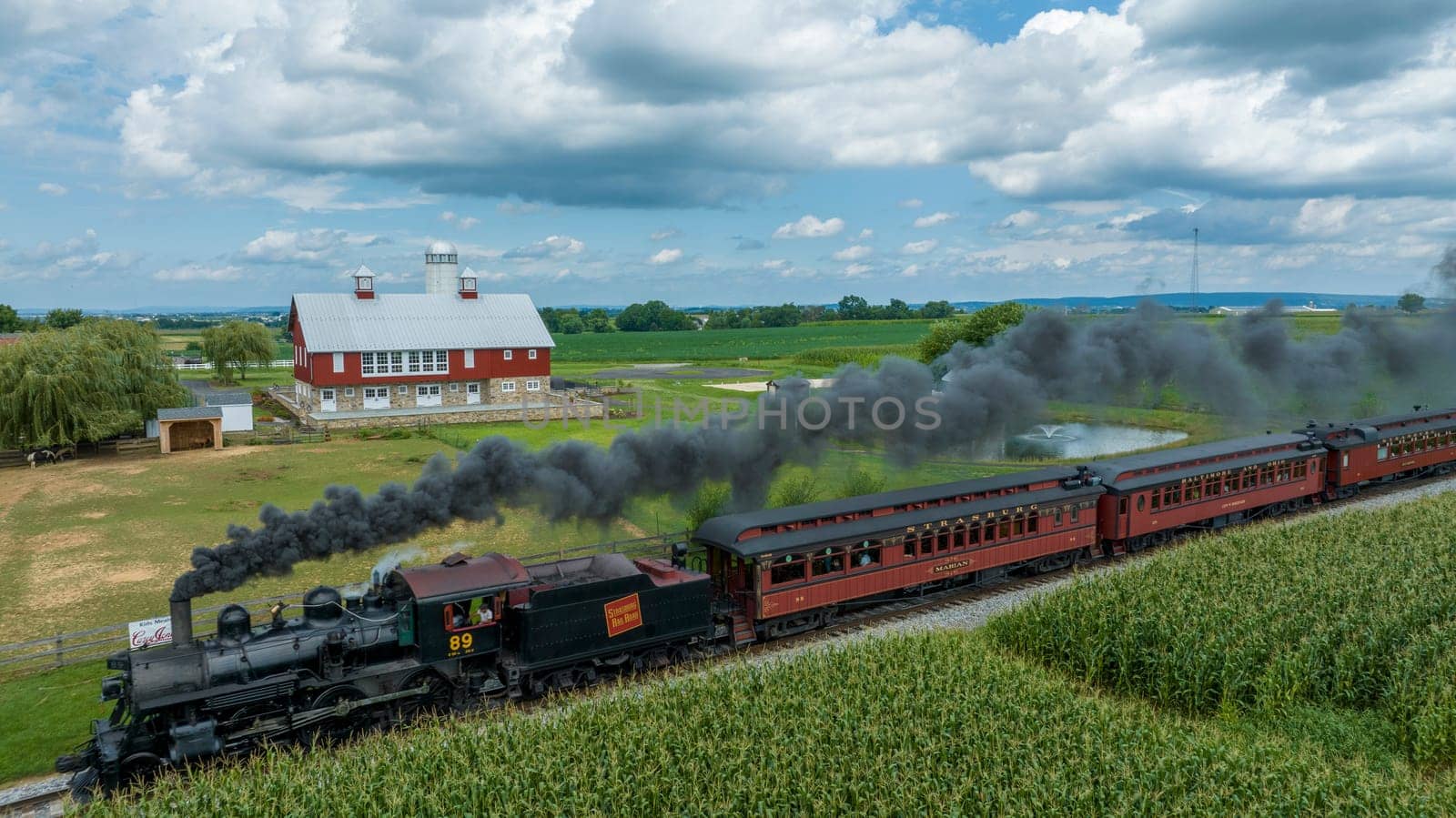 Ronks, Pennsylvania, August 15, 2023 - In a blend of motion and stillness, a steam train chugs along the tracks beside a tranquil farm, complete with a red barn and verdant fields.