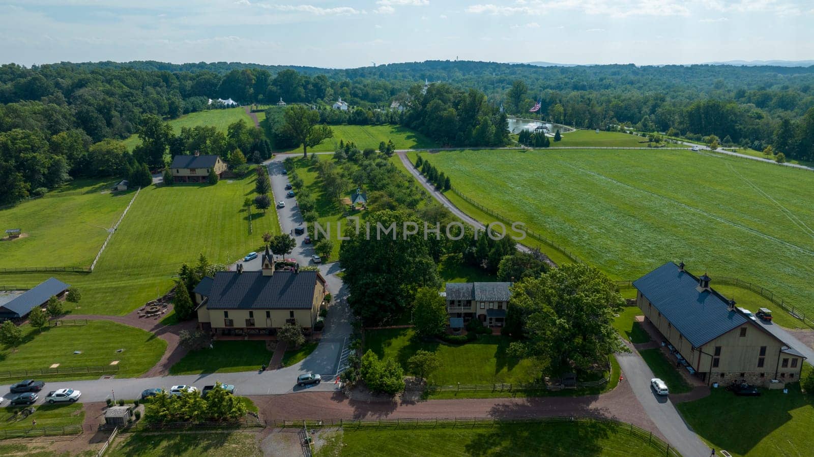 Overhead View Of A Serene Rural Landscape Featuring A Main Road Lined With Trees, Surrounded By Patches Of Green Fields, Fenced Areas, And A Collection Of Residential And Farm Buildings