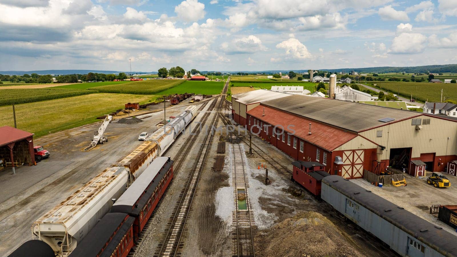 Strasburg, Pennsylvania, August 15, 2023 - An expansive view from above captures a rural rail yard with freight cars, surrounded by the lush, orderly farmlands stretching into the distance.