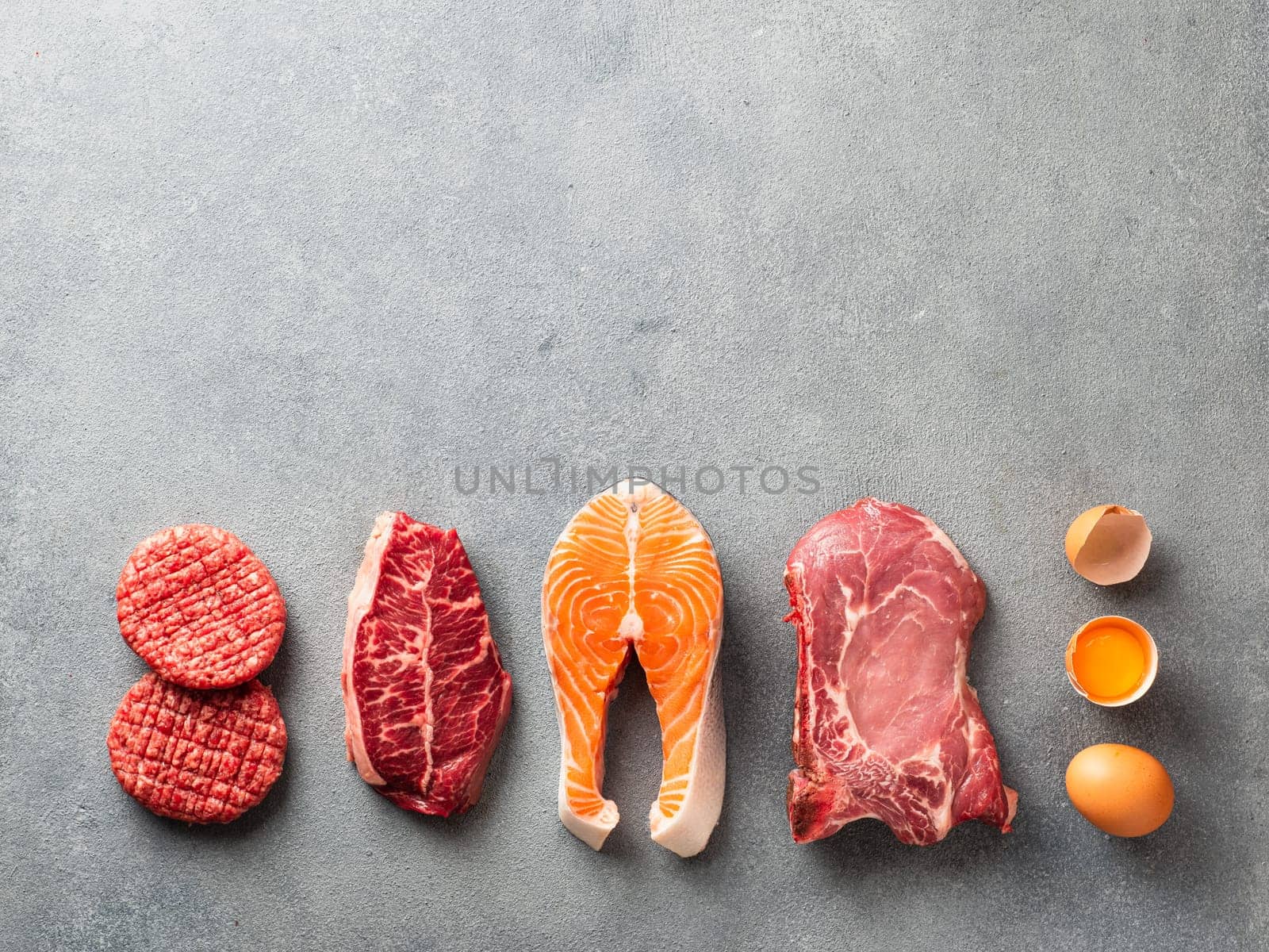 Carnivore or keto diet concept. Raw ingredients for zero carb or low carb diet - burger patties, ribeye, salmon steak, pork, egg on gray stone background. Top view or flat lay. Copy space top