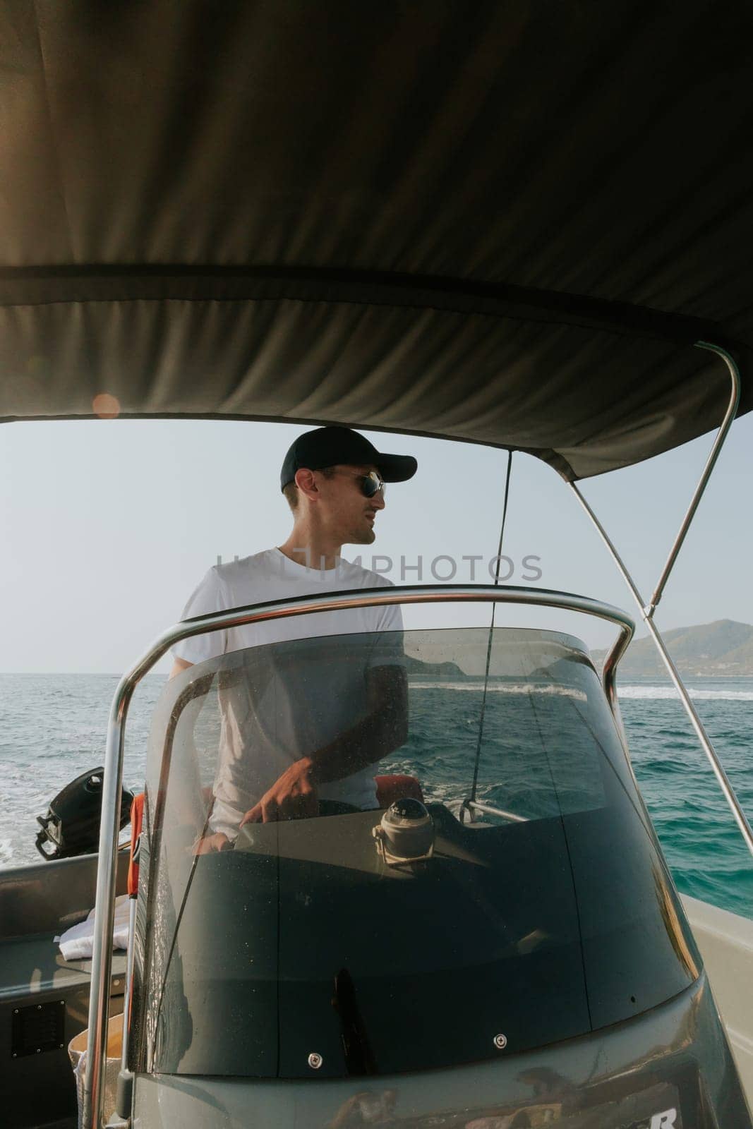 One young Caucasian guy in a cap and sunglasses sails on the sea on a boat, steering the helm and looking to the side, on a sunny summer day, close-up side view.