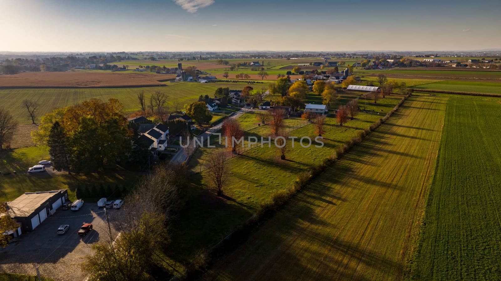 Aerial view of a sprawling farmland at sunset with long shadows, showcasing autumn's touch on trees and fields, perfect for themes of harvest, rural planning, and seasonal changes.