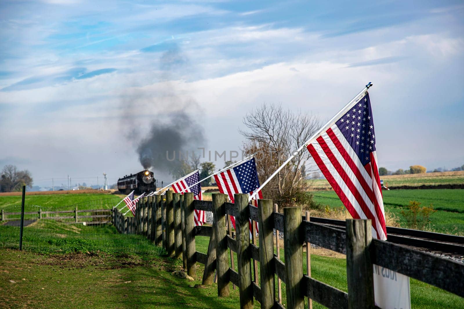 A vintage steam train chugs along the countryside, flanked by American flags on a wooden fence, evoking a sense of nostalgic Americana and historic travel.