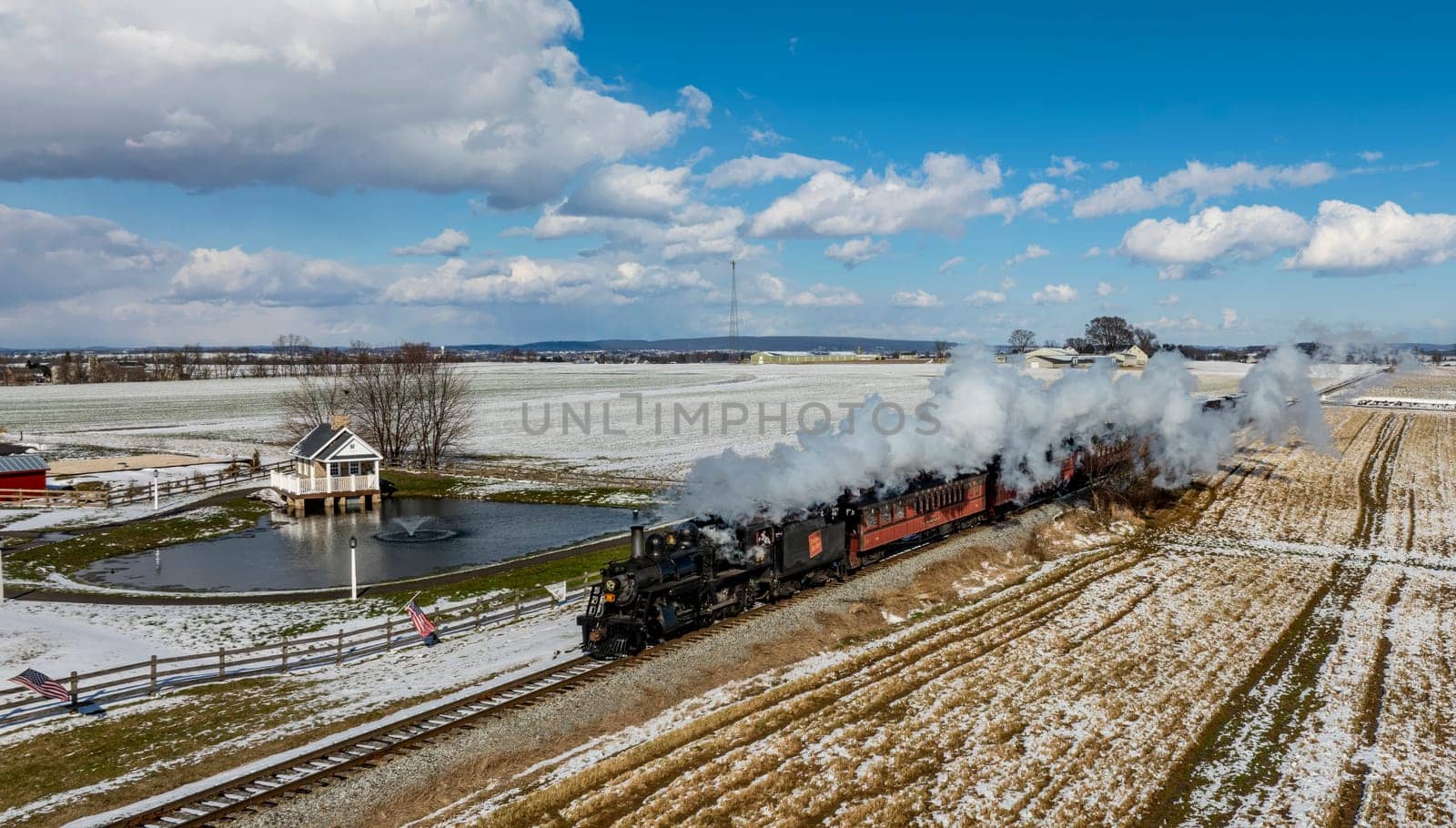 Strasburg, Pennsylvania, February 17, 2024 - A steam train chugs through a snow-dusted landscape, offering a scenic winter journey by the rails, with a quaint pond house nearby.