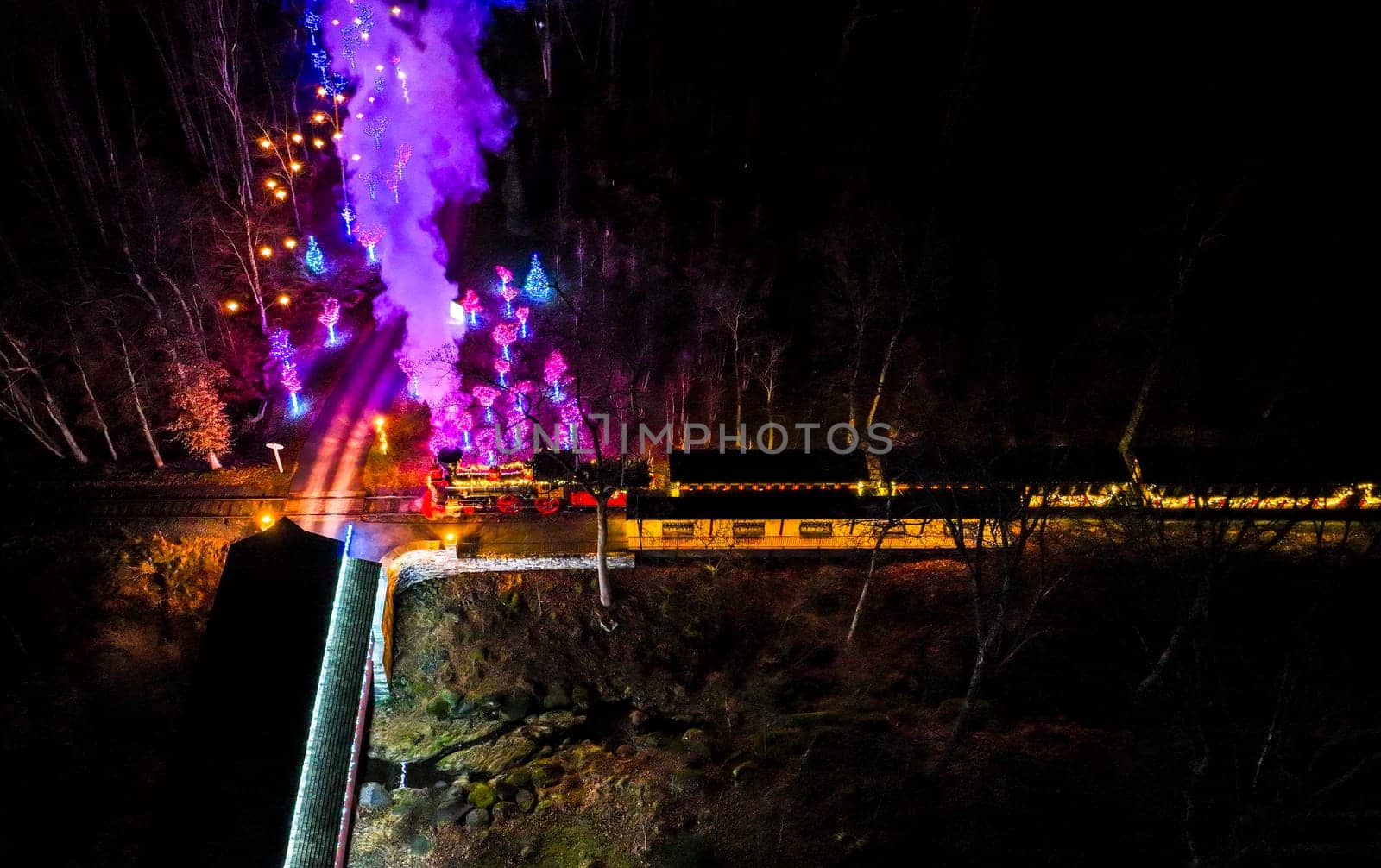 Aerial View Of A Vibrantly Lit Train Emitting Purple Smoke As It Passes Through A Forested Area At Night.