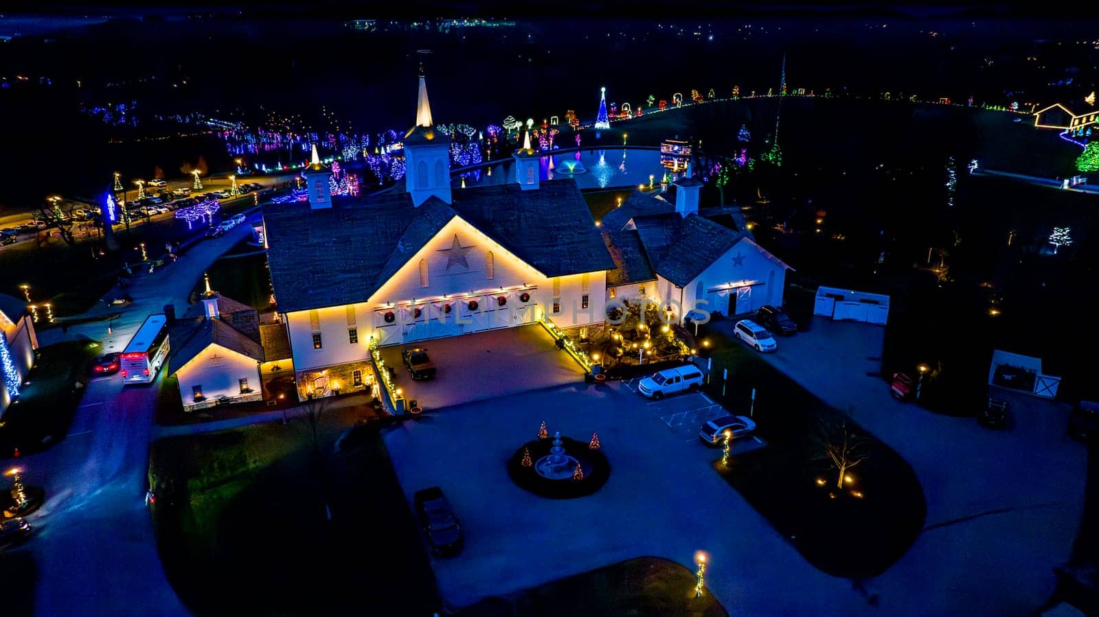 Overhead Night View Of A Complex With Buildings Outlined In Lights, A Central Pond, And A Surrounding Drive Lined With Lit Trees.