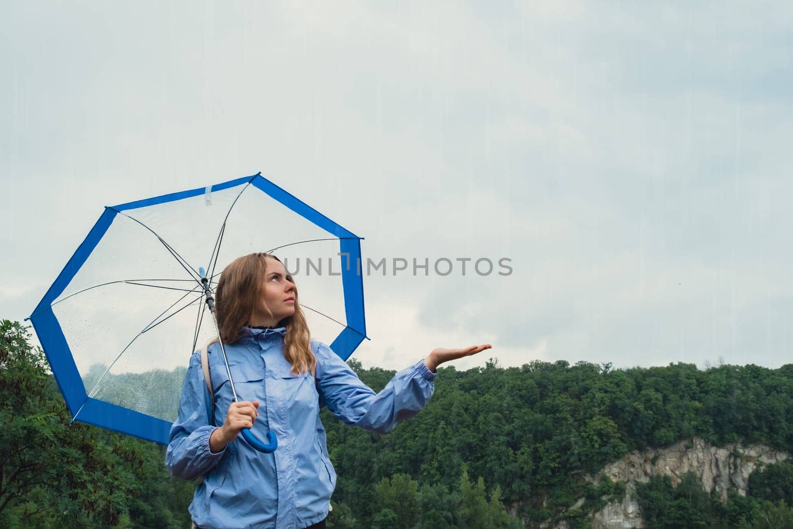 Young woman holding transparent blue umbrella outdoors in forest. Rainy weather day using umbrella. Woman with hand checking how long it will be raining by anna_stasiia