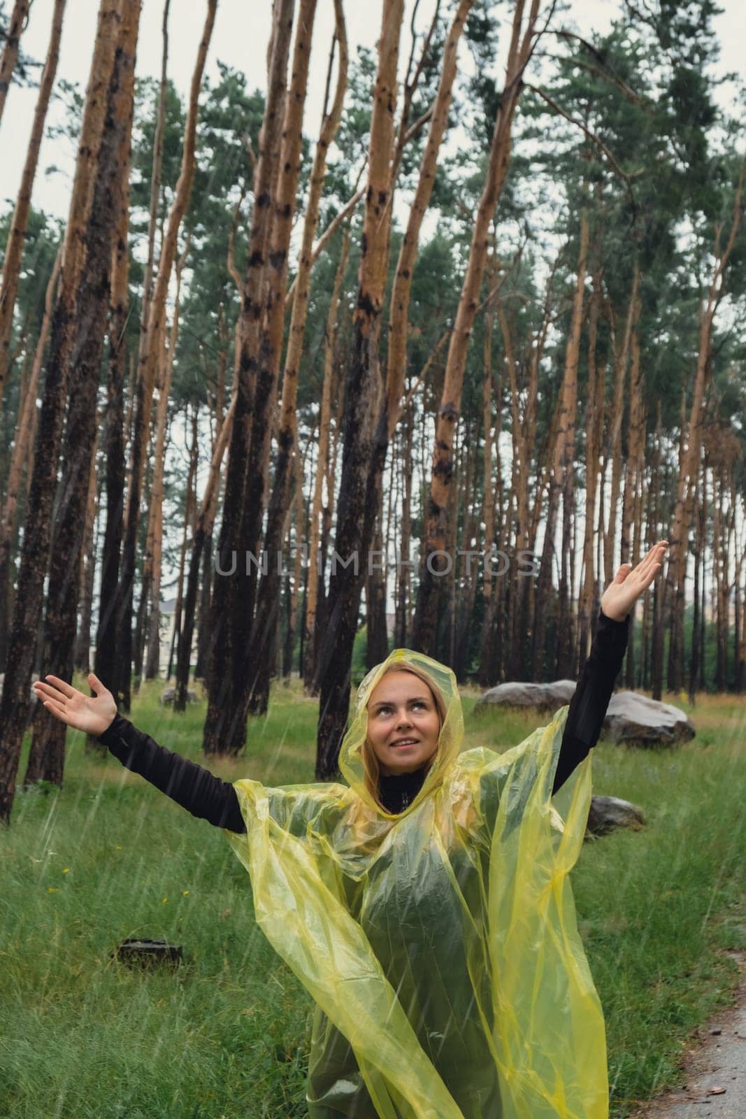 Happy blonde woman in yellow raincoat enjoying outdoors in forest. Rainy weather outside leisure activities in autumn time. Rainy season forecast